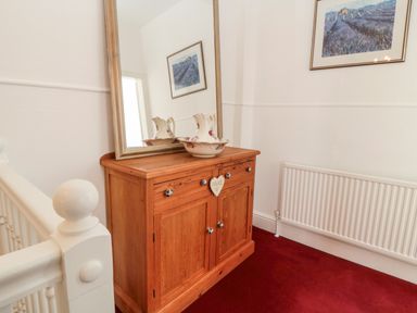 A wooden cabinet with a mirror and floral ceramic pitcher on top in a hallway with red carpet at The Robins in Filey