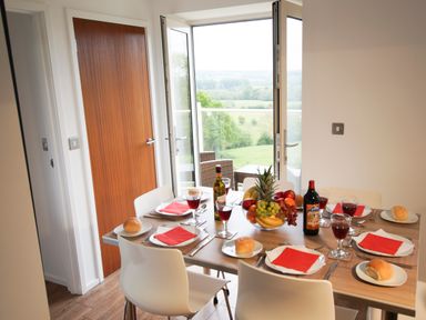 A dining table set with plates bread red napkins and glasses near open glass doors overlooking a landscape at Beautiful Cotswold View in Bourton-On-The-Water
