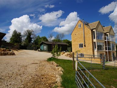 A gravel driveway with a green cabin and a stone house with large windows and a metal gate at Beautiful Cotswold View in Bourton-On-The-Water
