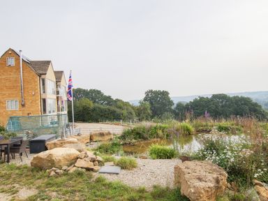 A garden with rocks and plants next to a pond and a building with British flags at Beautiful Cotswold View in Bourton-On-The-Water