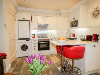 A kitchen with white cabinets a washing machine red bar stools and purple flowers in a vase at Church View in Ambleside