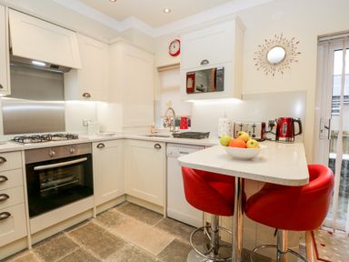 A kitchen with white cabinets and countertops a gas stove oven sink microwave red kettle toaster and two red bar stools at Church View in Ambleside