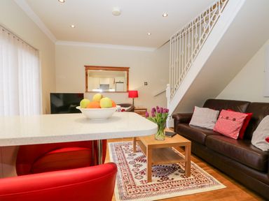 A living room with a brown leather sofa red and gray cushions a wooden coffee table with a vase of purple flowers a patterned rug a white countertop bowl of fruit and a mirror on the wall at Church View in Ambleside