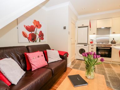 A living area with a brown leather sofa with cushions and a wooden table with a vase of purple flowers and a closed book in a kitchen space at Church View in Ambleside