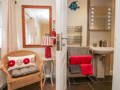 A wicker chair with cushions next to a small table with a lamp and a mirror on the wall beside a bathroom with a sink and towel rack at Church View in Ambleside