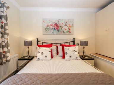A bedroom with a bed with striped bedding and cushions with sheep patterns and red pillows two bedside tables with lamps and floral curtains at Church View in Ambleside