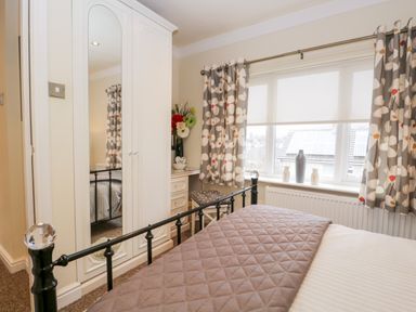 A bedroom with a bed a white wardrobe with mirrored doors a window with patterned curtains and a small dressing table with flowers at Church View in Ambleside