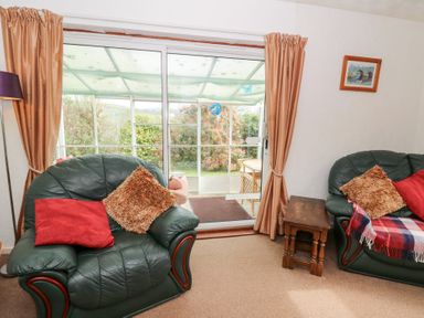 A living room with green leather armchairs with cushions and a small wooden table near sliding glass doors opening to a garden at Bryn yr Haf in Llanfair Clydogau near Lampeter