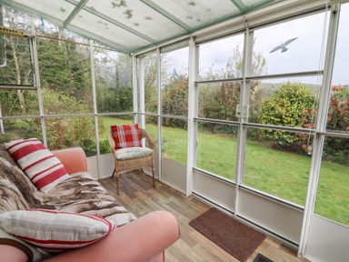 A sunroom with a sofa, cushion, wicker chair, and a view of a garden at Bryn yr Haf in Llanfair Clydogau near Lampeter