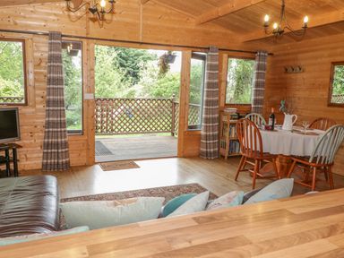 A wooden room with a dining table set with chairs next to windows and glass doors opening to a deck outside at Y Caban in Llechwedd The Conwy Valley
