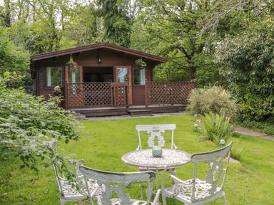 A wooden cabin with a porch and lattice railing in a garden with a white metal table and chairs surrounded by trees at Y Caban in Llechwedd The Conwy Valley