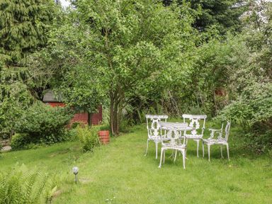 A garden with white metal chairs and table on grass surrounded by trees and bushes at Y Caban in Llechwedd The Conwy Valley