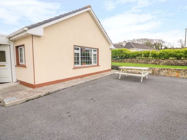 An exterior view of a cream colored building with a window and a door next to a paved area with a white picnic table and green hedges at Island View in Ardmore County Waterford