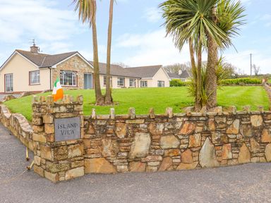 A stone wall with an island view sign and an Irish flag in front of a house with palm trees at Island View in Ardmore County Waterford