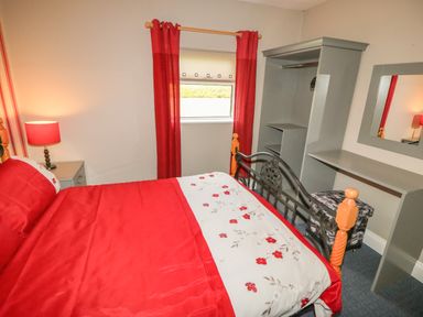 A bedroom with a bed covered in red and white bedding with red curtains on the window and a gray wardrobe desk and mirror at Island View in Ardmore County Waterford