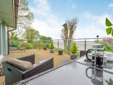 A wooden deck with wicker chairs and a glass table with a coffee press overlooking a glass railing and a coastal view at Solway Cottage in Bowness-on-Solway near Wigton