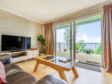 A living room with a brown leather sofa a wooden coffee table a flat screen TV on a stand and a sliding glass door leading to a deck overlooking water at Solway Cottage in Bowness-on-Solway near Wigton
