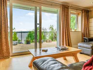 A living room with a wooden coffee table and leather chairs next to glass sliding doors leading to a balcony with potted plants at Solway Cottage in Bowness-on-Solway near Wigton