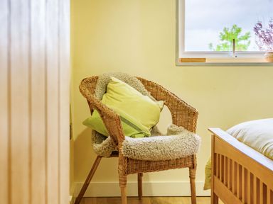 A wicker chair with cushions and a sheepskin throw next to a bed and a window showing trees at Solway Cottage in Bowness-on-Solway near Wigton