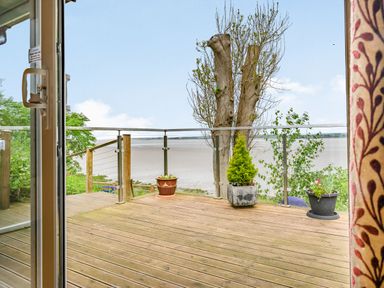 A wooden deck with potted plants and glass railing overlooking a body of water at Solway Cottage in Bowness-on-Solway near Wigton