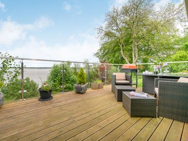 A wooden deck with outdoor wicker furniture a barbecue and potted plants overlooking a body of water at Solway Cottage in Bowness-on-Solway near Wigton