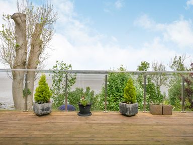A wooden deck with potted plants and glass railing overlooking a beach and trees at Solway Cottage in Bowness-on-Solway near Wigton