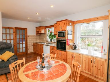A kitchen with wooden cabinets, a tiled round dining table with chairs, a window with plants, and an oven at Raheen Castletownshend County Cork
