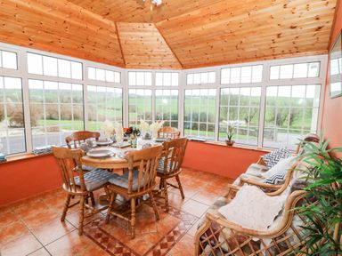 A sunroom with a wooden dining table and chairs set for a meal and a wicker sofa with cushions at Raheen in Castletownshend County Cork