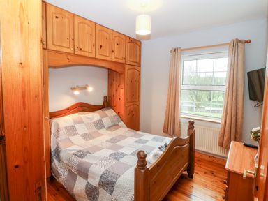 A bedroom with a wooden bed framed by built-in wooden cabinets a window with curtains and a wooden dresser at Raheen in Castletownshend County Cork