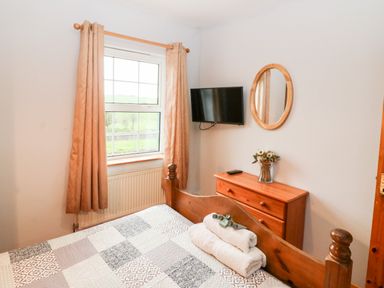A bedroom with a bed and folded towels a wooden dresser with a vase of flowers a round mirror and a wall-mounted TV at Raheen in Castletownshend County Cork