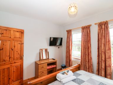 A bedroom with a wooden door wooden dresser with mirror mounted TV two windows with patterned curtains and a bed with folded towels at Raheen in Castletownshend County Cork
