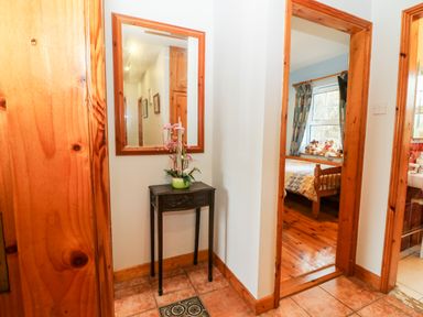 A hallway with a small black table and a mirror above it and doors leading to a bedroom with stuffed animals on the bed and a bathroom at Raheen Castletownshend County Cork