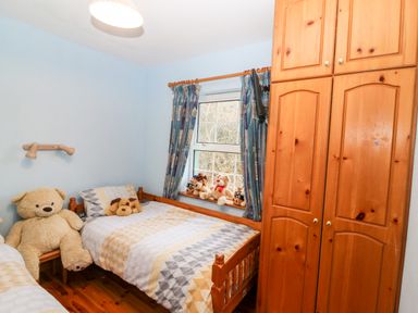 A bedroom with two wooden beds with patterned bedding a large teddy bear on a chair stuffed animals on the window sill blue patterned curtains and a tall wooden wardrobe at Raheen Castletownshend County Cork