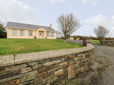 A single-story house with multiple windows lawn with two leafless trees stone wall and gated driveway at Raheen in Castletownshend County Cork
