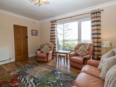 A living room with leather armchairs a sofa a colorful rug wooden tables and large window with striped curtains at Bron Aber in Arthog near Dolgellau