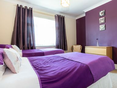 A bedroom with two beds covered in purple bedspreads a wooden chest of drawers a wicker chair and purple curtains at Tomeg in Amble-by-the-Sea