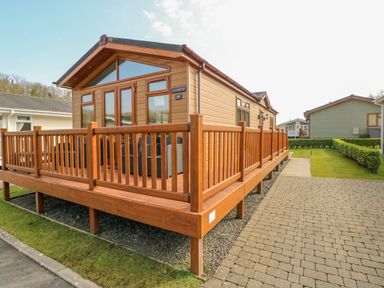 A wooden cabin with a fenced deck and outdoor chairs on a paved and grassy area at Cladebrine Wisemans Bridge near Saundersfoot