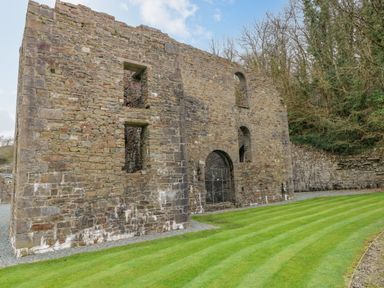 An exterior view of a stone ruin with arched windows and doorways next to a striped grass lawn and trees at Cladebrine Wisemans Bridge near Saundersfoot