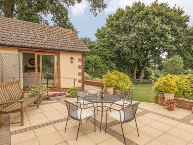 An outdoor patio with a metal table and four chairs a wooden bench potted plants and a sliding glass door leading inside at Pipistrelle Lodge in Plymtree