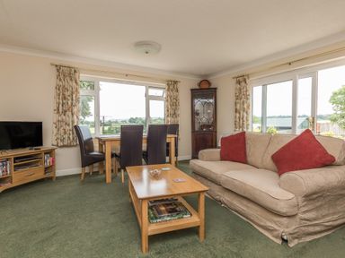 A living room with a beige sofa with red cushions a wooden coffee table a wooden dining table with four black chairs a television on a wooden stand and large windows with floral curtains at Pipistrelle Lodge in Plymtree