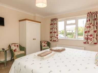 A bedroom with a bed, wardrobe, two chairs, side table, and window with floral curtains at Pipistrelle Lodge in Plymtree
