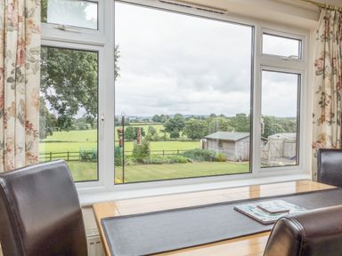 A dining table with leather chairs next to a large window showing a garden and green fields at Pipistrelle Lodge in Plymtree