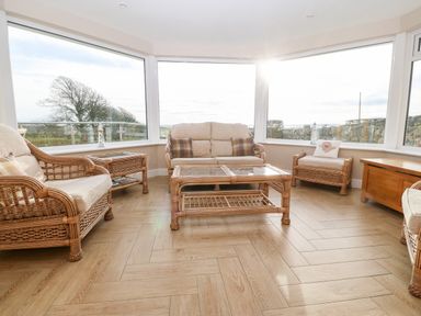 A sunroom with rattan furniture and large windows showing an outdoor view at Ty Top in Llanfaethlu