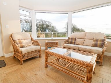 A seating area with wicker armchair and sofa and glass top coffee table in a room with large windows at Ty Top in Llanfaethlu