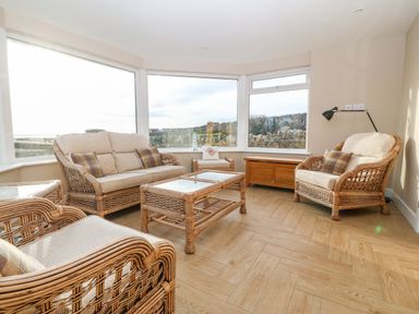 A sitting room with wicker furniture and large windows showing an outdoor view at Ty Top in Llanfaethlu