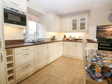 A kitchen with cream cabinets a brown countertop a sink a stove and a table with a pebble patterned tablecloth at Ty Top in Llanfaethlu