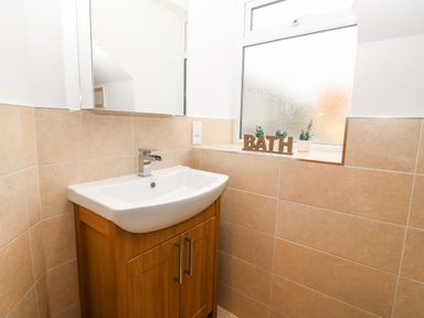 A bathroom sink with wooden cabinet below a mirror next to a frosted window with bath letters and small plants on the ledge at Ty Top in Llanfaethlu