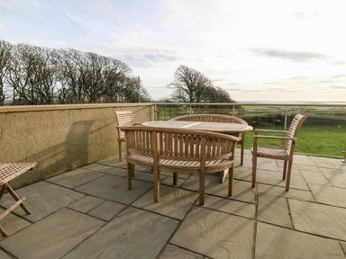 An outdoor patio with wooden chairs and a wooden table overlooking fields and trees at Ty Top in Llanfaethlu