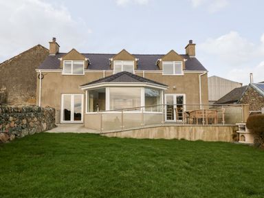 A two-story house with bay windows and a patio with wooden furniture and glass railing in front of a lawn at Ty Top in Llanfaethlu