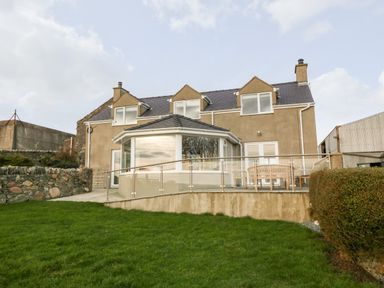 The exterior of a two-story house with a grass lawn and a patio with a wooden bench at Ty Top in Llanfaethlu